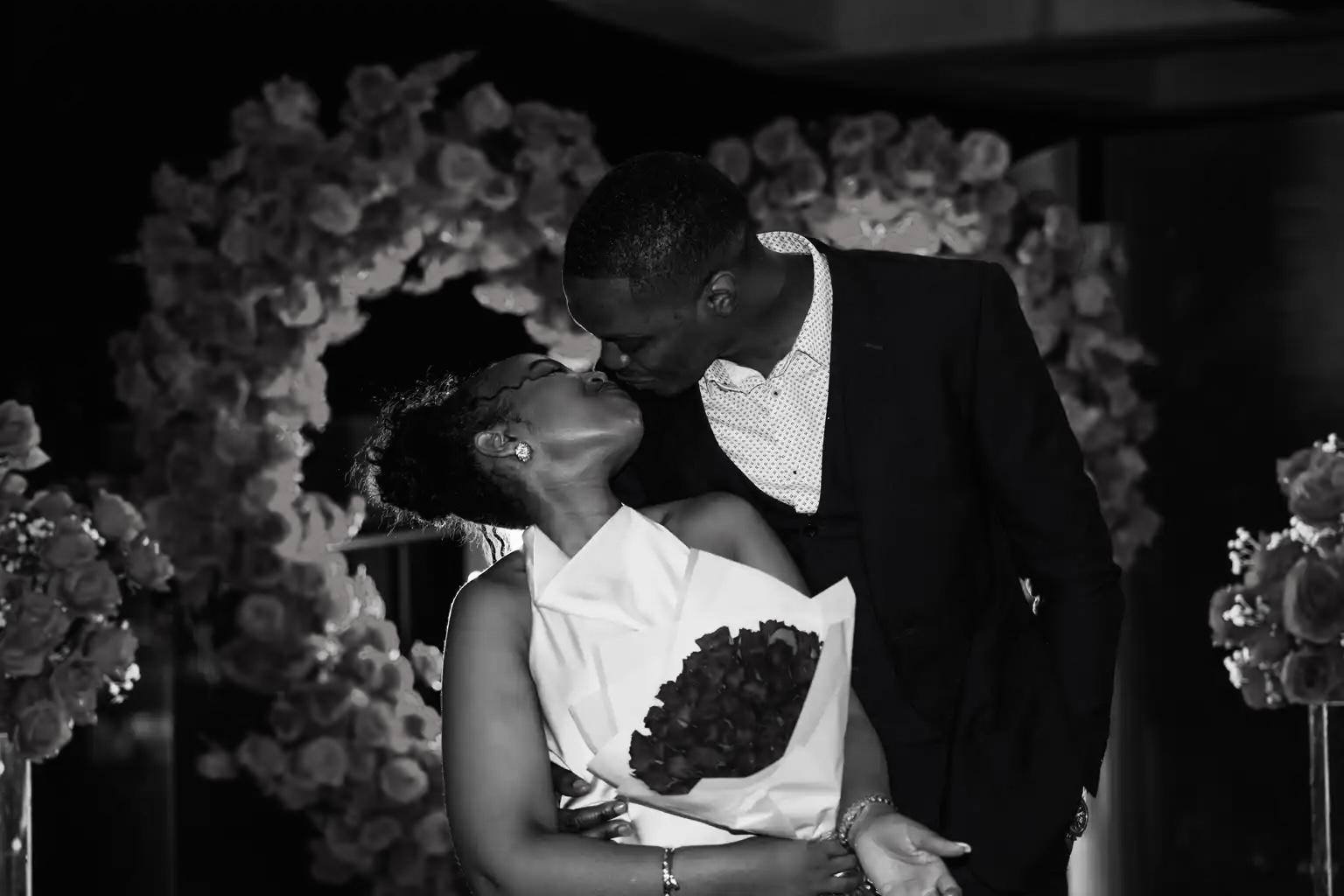 Afifoluwa and Mihlali kissing in front of a heart-shaped floral arch, holding a bouquet of roses, in black and white