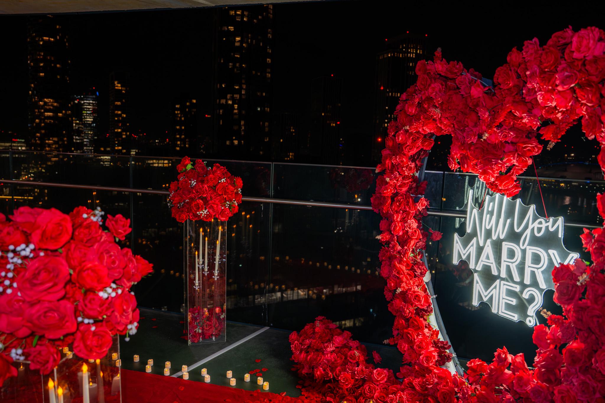 The proposal setting at night: a heart-shaped arch of red roses framing a neon Will You Marry Me? sign, with candles and rose arrangements above the city skyline