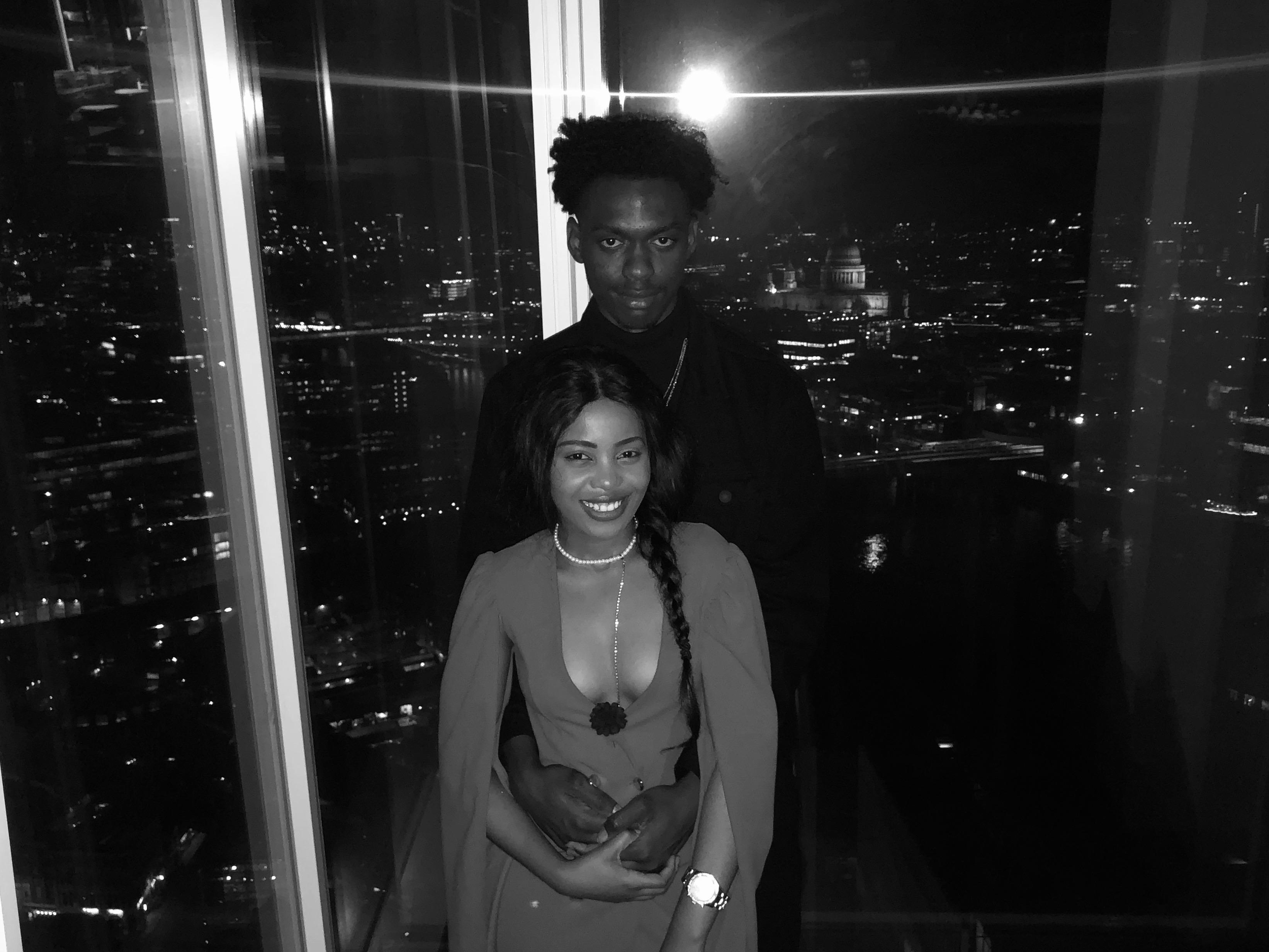 Afifoluwa and Mihlali at a window high above London at night, the lit city and St Paul's behind them, in black and white